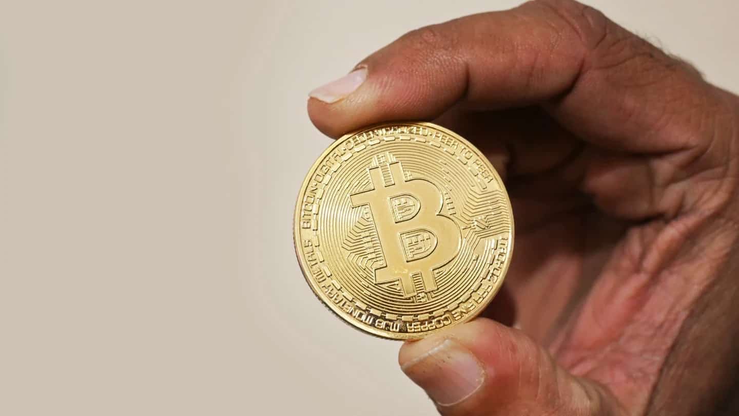 Mão segurando uma moeda dourada com o símbolo do bitcoin, contra fundo claro.