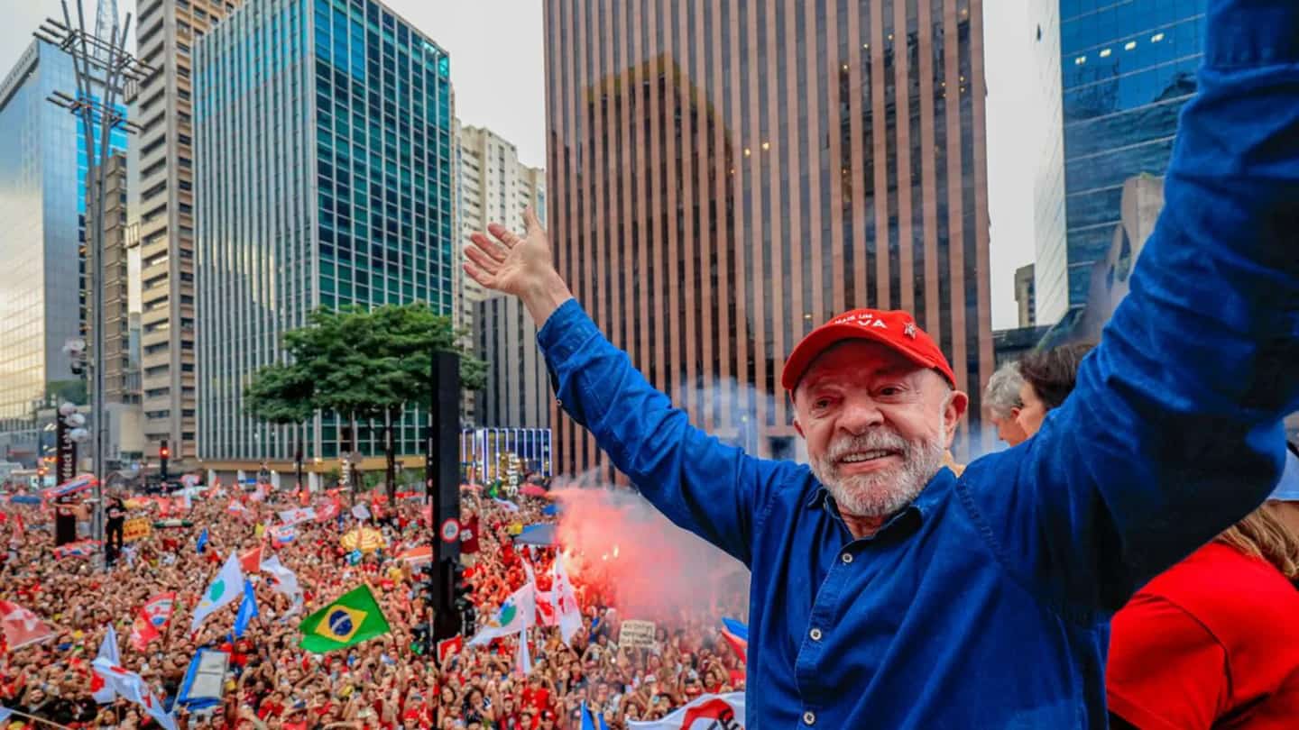 Lula ergue os braços para a multidão durante ato público, com bandeiras do Brasil e faixas de campanha ao fundo