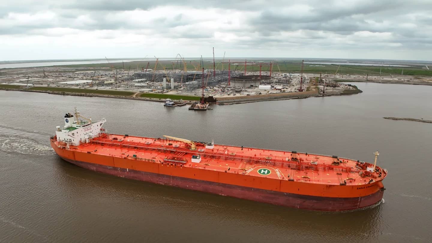 Navio petroleiro carregado de petróleo bruto navegando em canal portuário, com instalações industriais ao fundo
